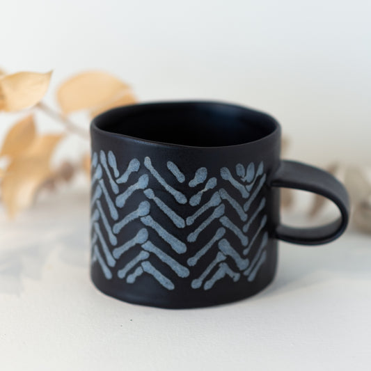 Black mug with white chevron pattern in front of a yellow plant.