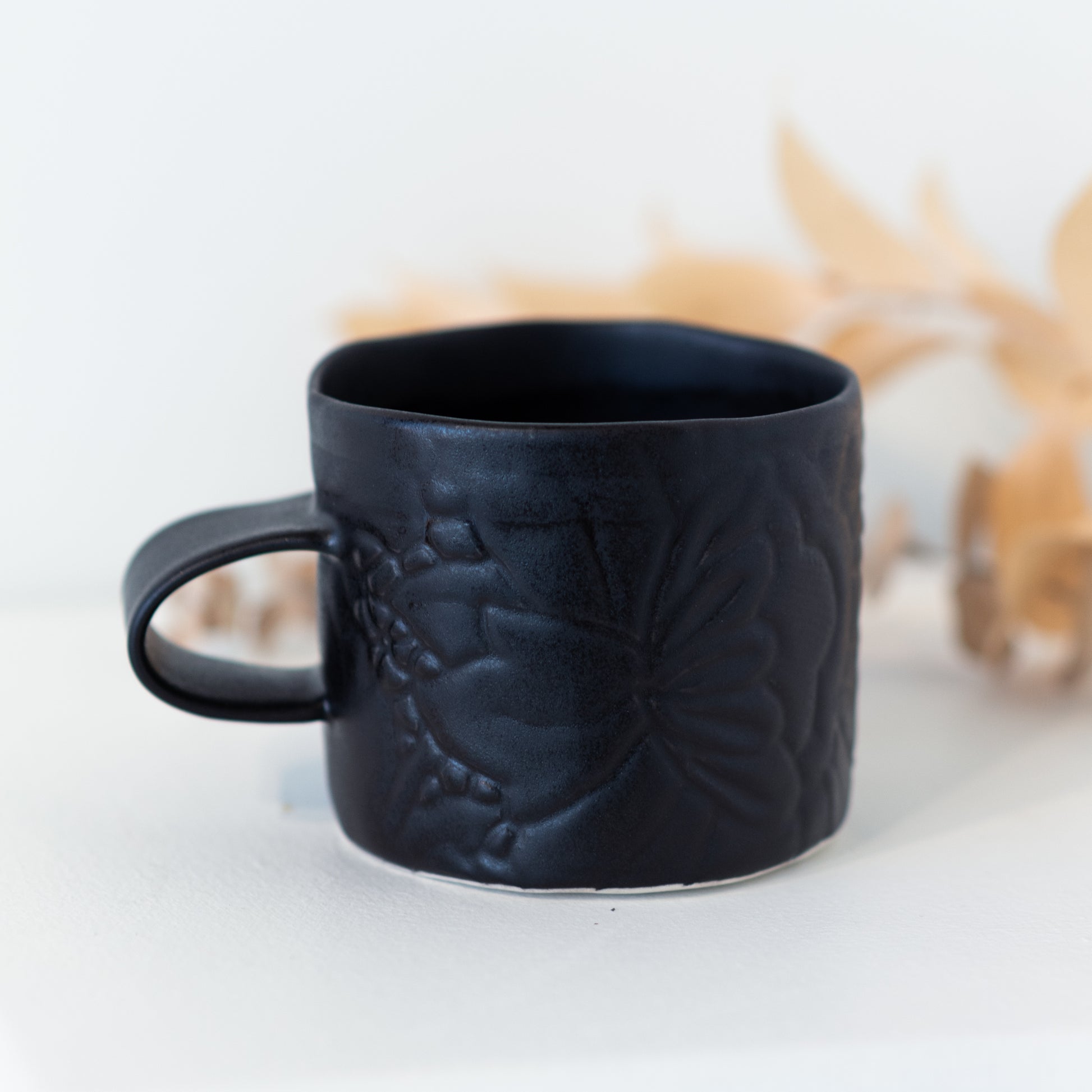 Black mug with subtle floral patterning in front of a yellow plant.