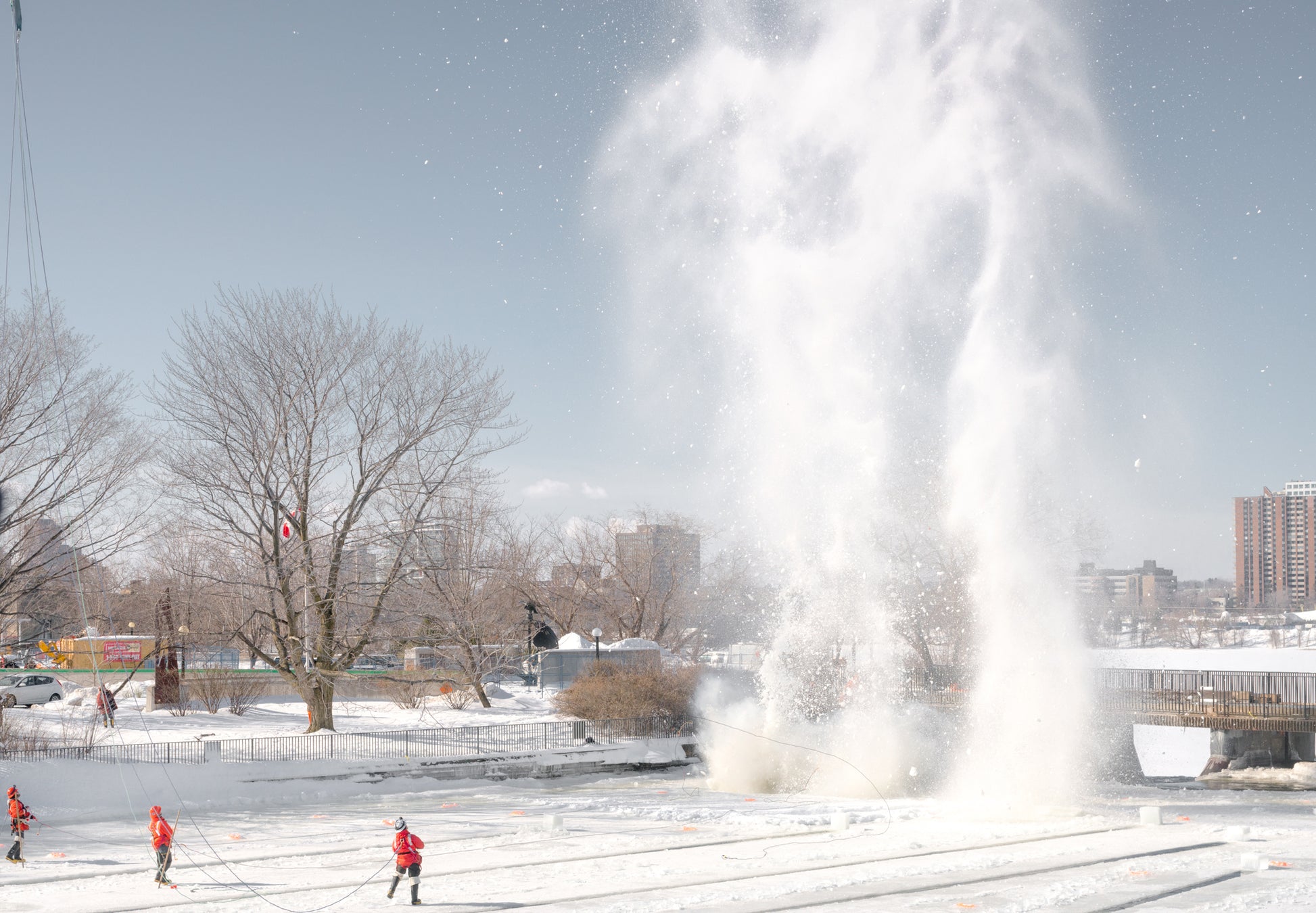 Three men in orange coats in front of a blast of snow that rises up into the sky above the trees.