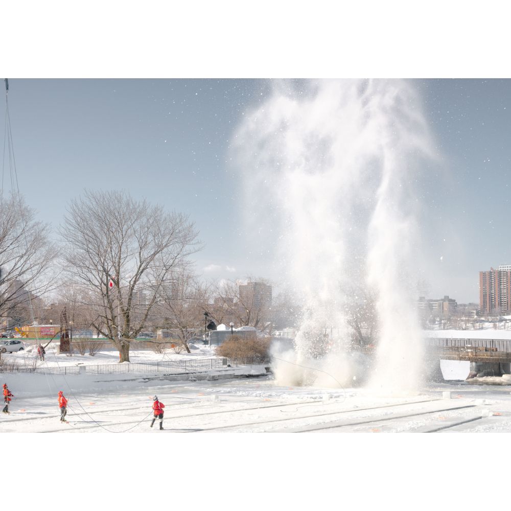 Three men in orange coats in front of a blast of snow that rises up into the sky above the trees.